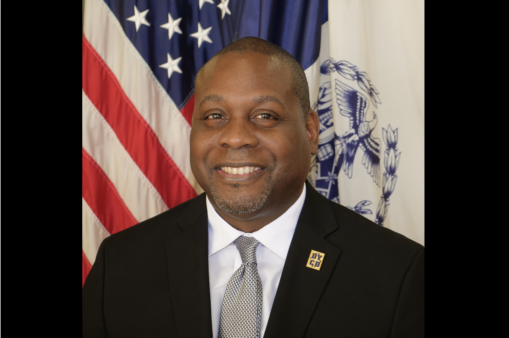 A man is wearing a suit and smiling in front of the American Flag